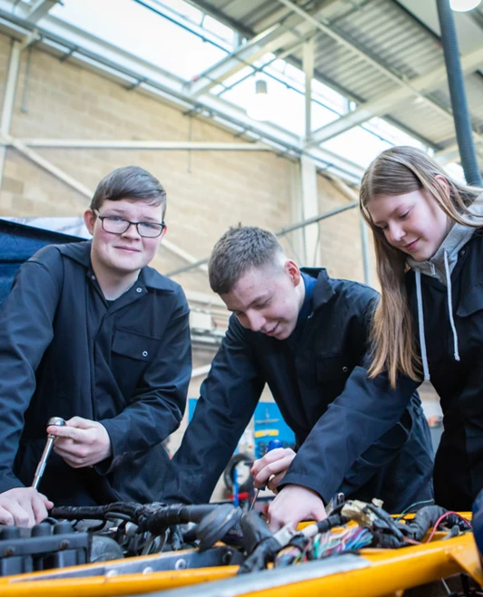 Three students working together under the hood of a car, using tools to inspect and repair an engine. Three students working together under the hood of a car, using tools to inspect and repair an engine.